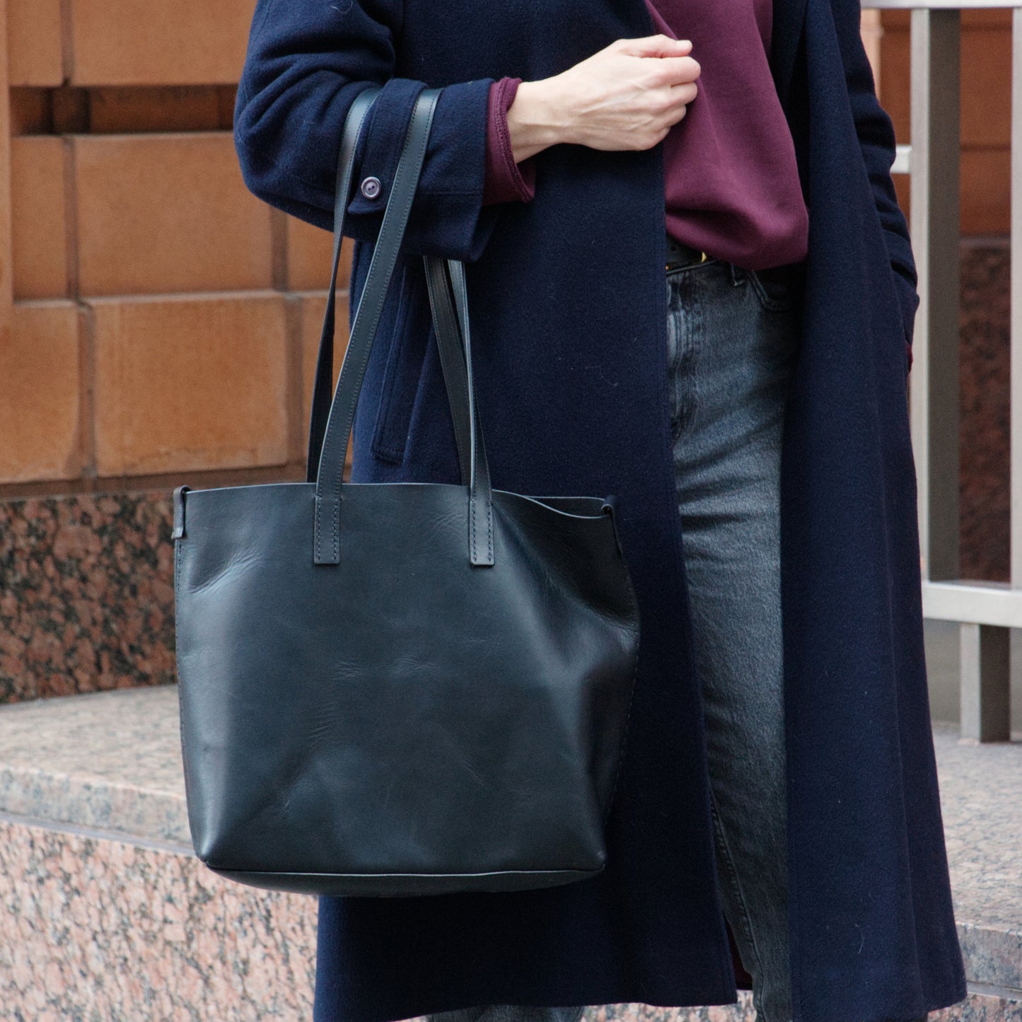 Person holding a black leather tote bag against a brick wall.