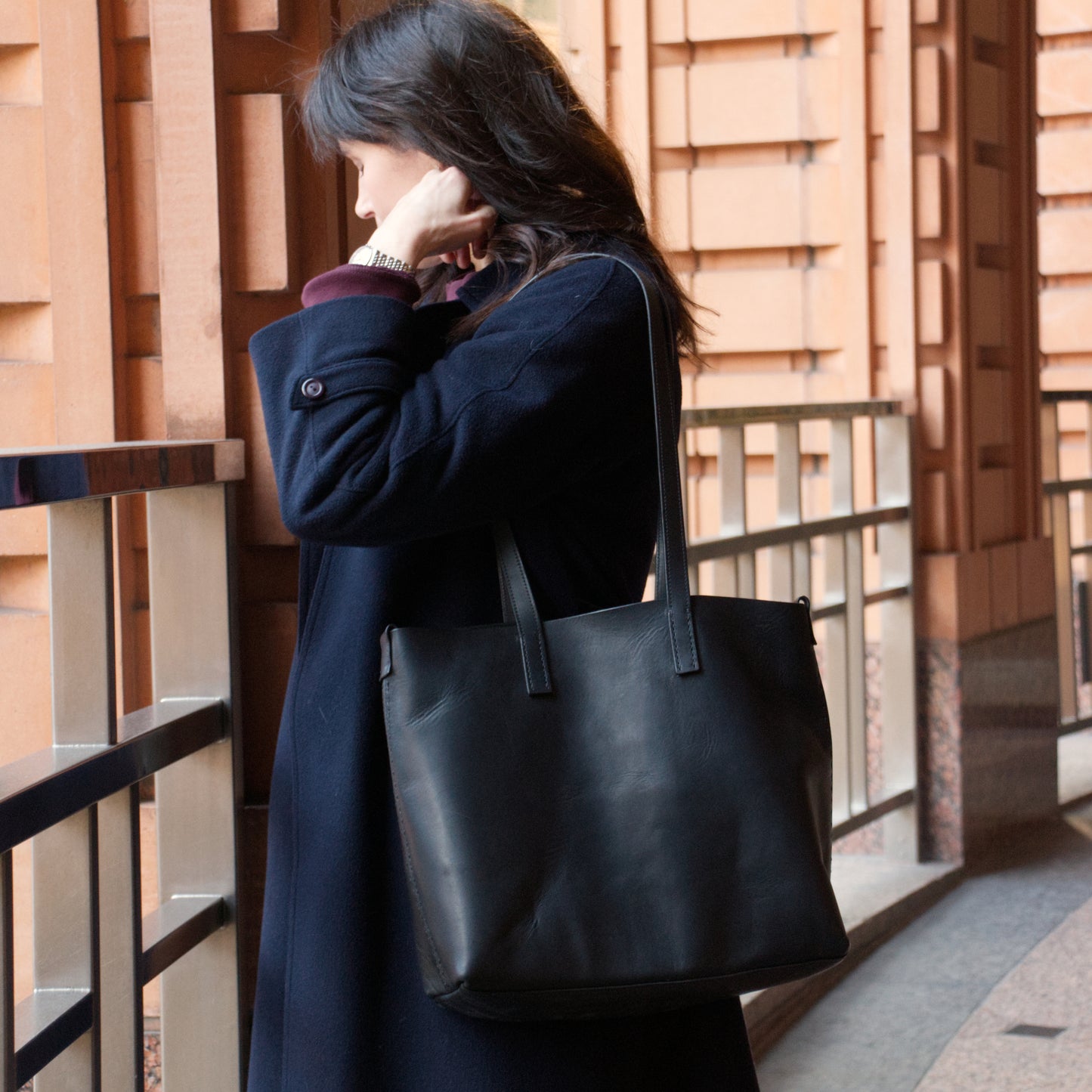 Person wearing a dark coat and holding a black leather bag against a wooden wall.